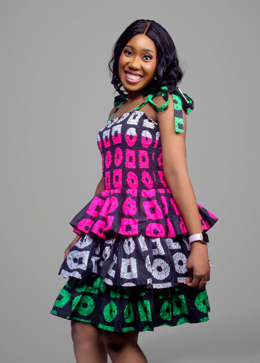 Front view of a smocked-bodice tiered dress in a pink, white, and green geometric African print with tie shoulders.