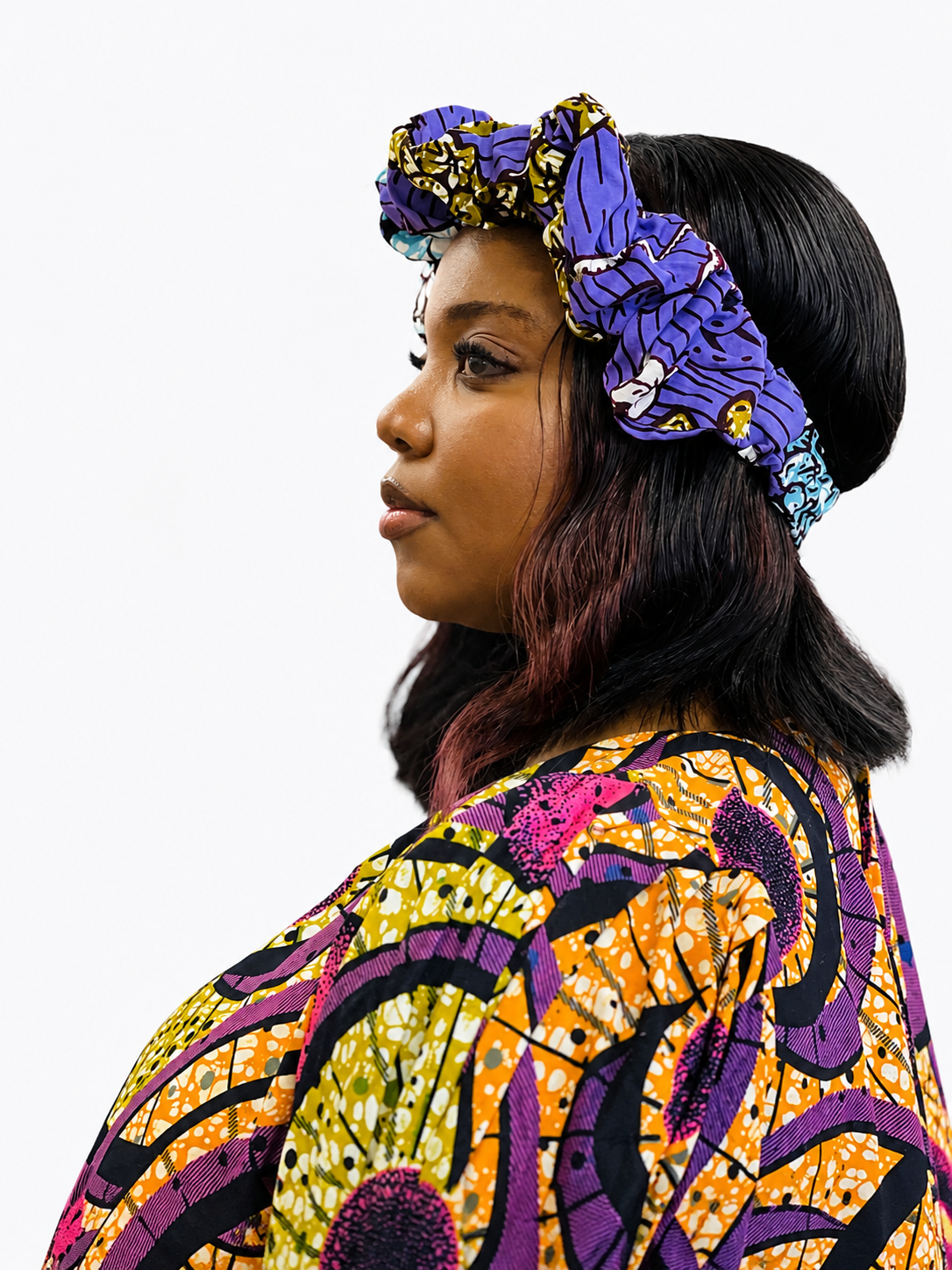 Side profile view of a person wearing a vibrant purple and orange ruffled African print headband made of 100% cotton.
