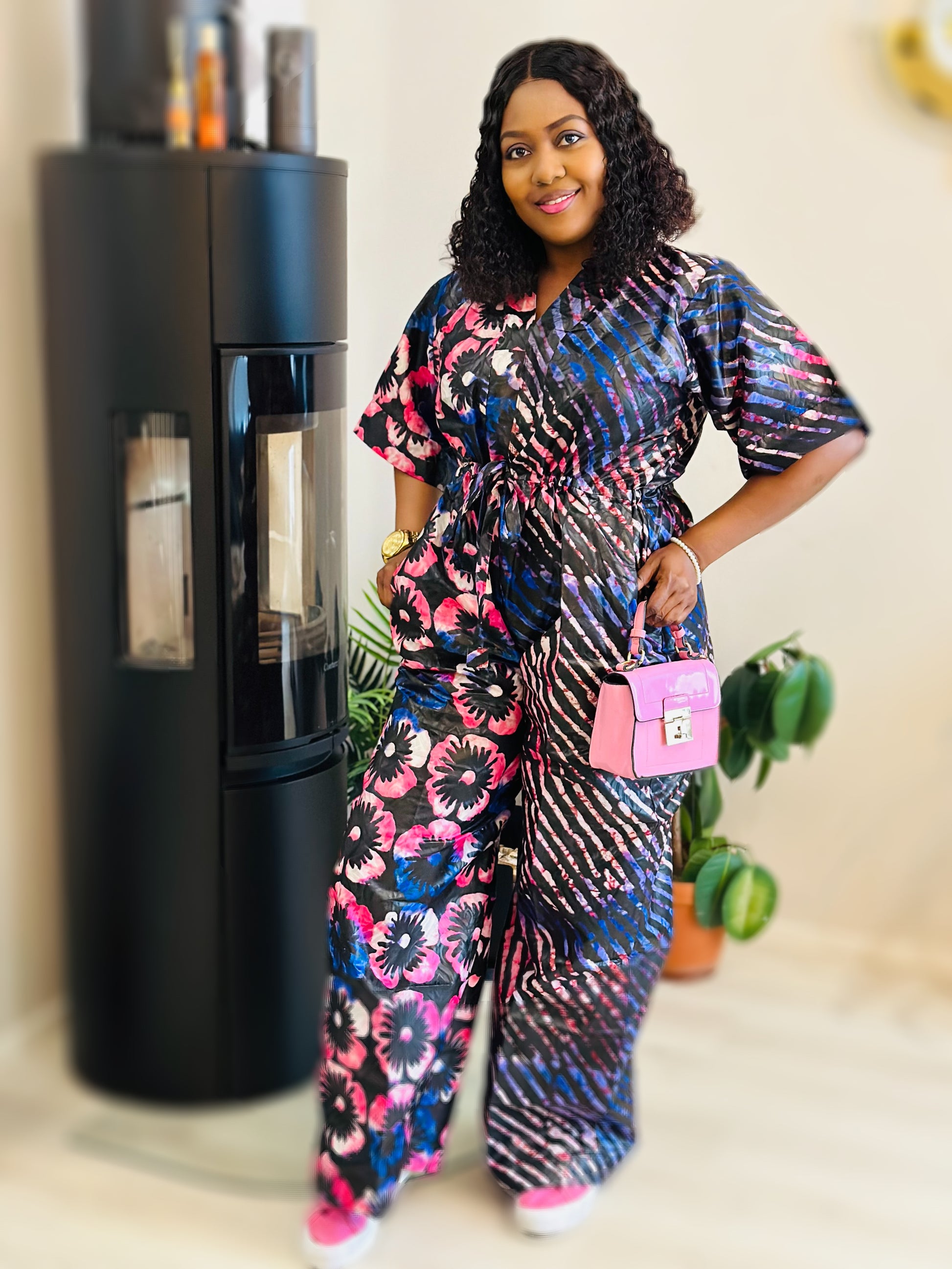 Woman posing indoors wearing a shiny floral and striped jumpsuit with a pink purse.