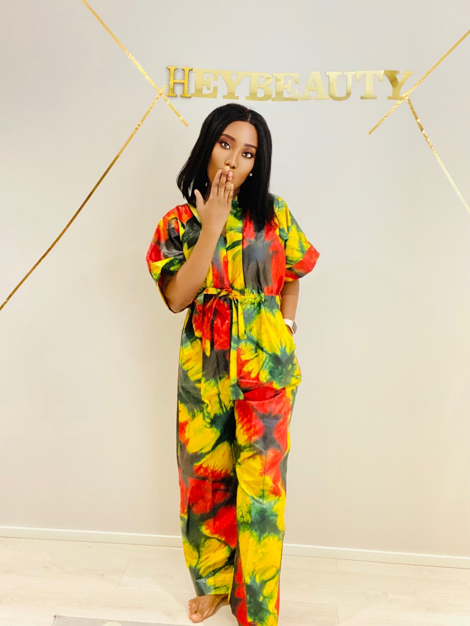 Barefoot woman posing in a colorful yellow, red, and green tie dye jumpsuit.